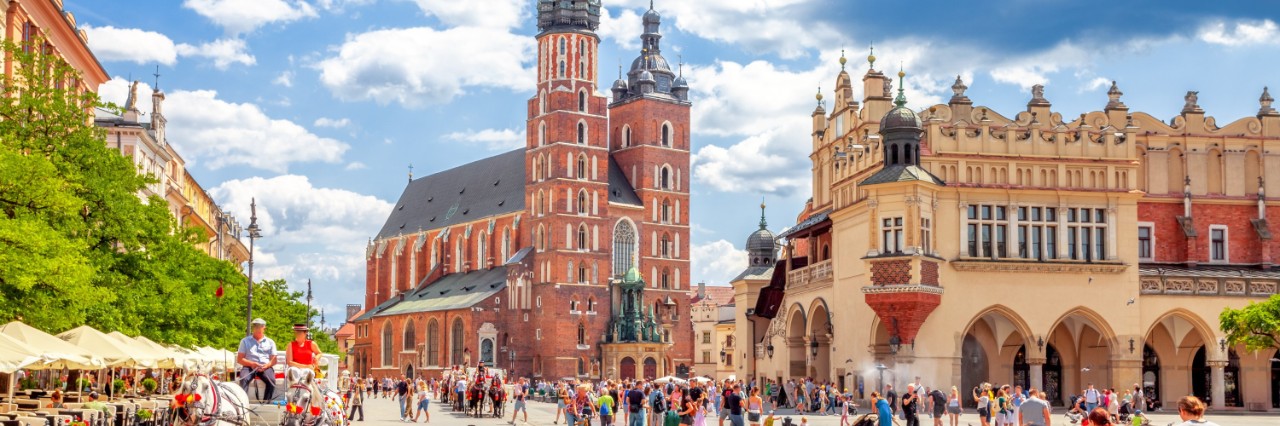 View of the Main Square in Krakow. On the left is a horse-drawn carriage, on the square several people. In the centre of the picture is St. Mary’s Basilica, to the right of it the Cloth Hall © Sina Ettmer/stock.adobe.com