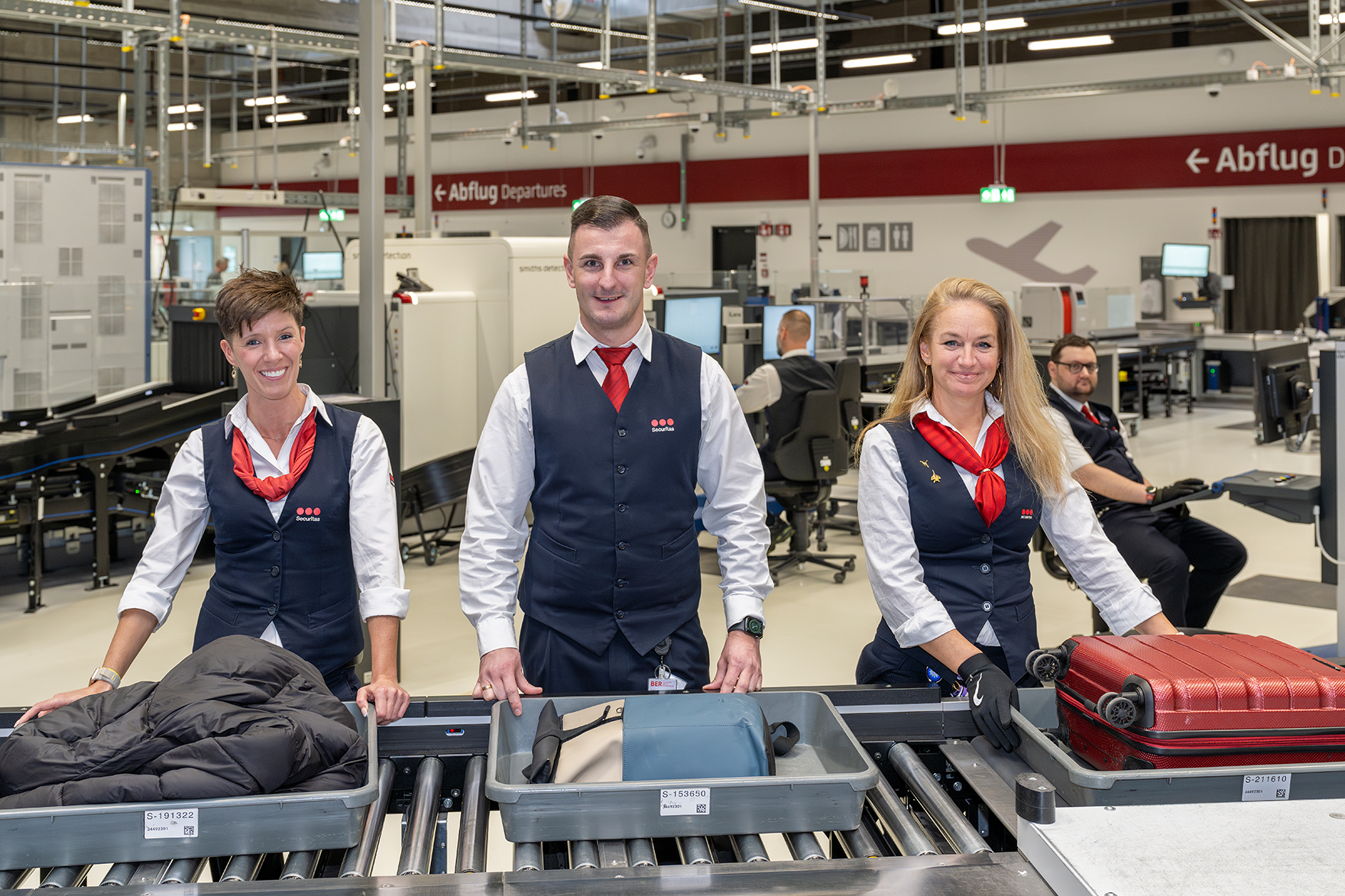 3 Securitas employees look into the camera at the conveyor belt. Luggage tubs in front of them.