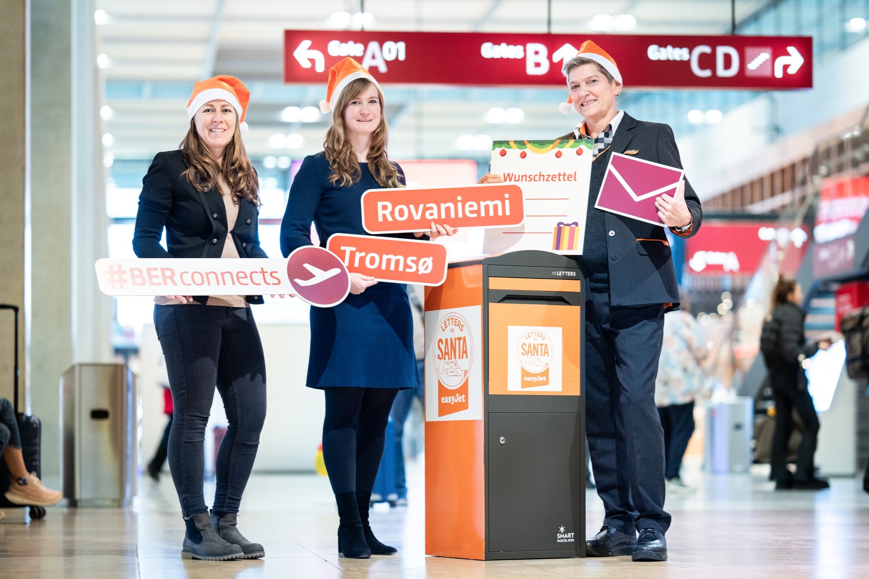 Three people are standing in the terminal wearing Santa hats and holding up signs. There is a large letterbox between them.