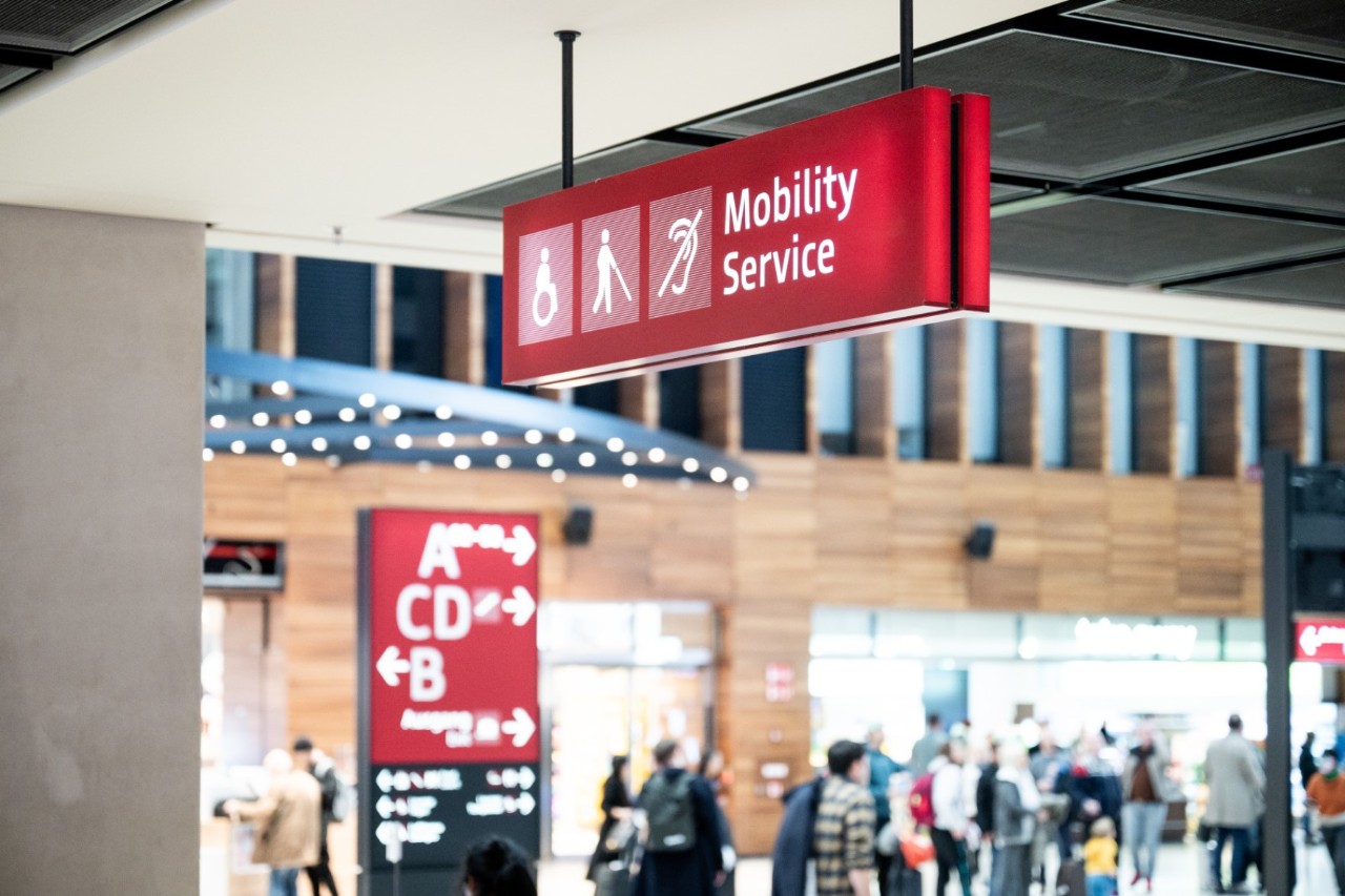 Mobility Service © Anikka Bauer / FBB Schild mit der Aufschrift Mobility Service. Dahinter Menschen im Marktplatz.