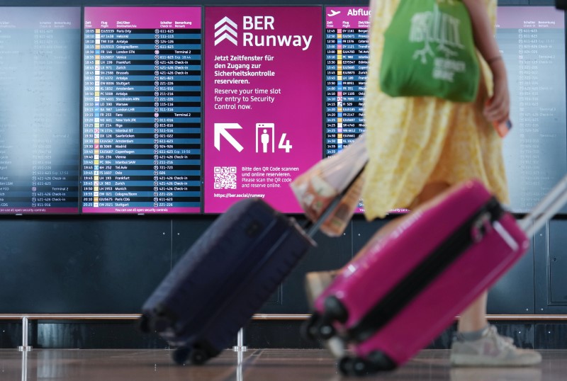 A person pulls a suitcase through the picture. In the background is the flight board and a monitor with a sign for BER Runway.
