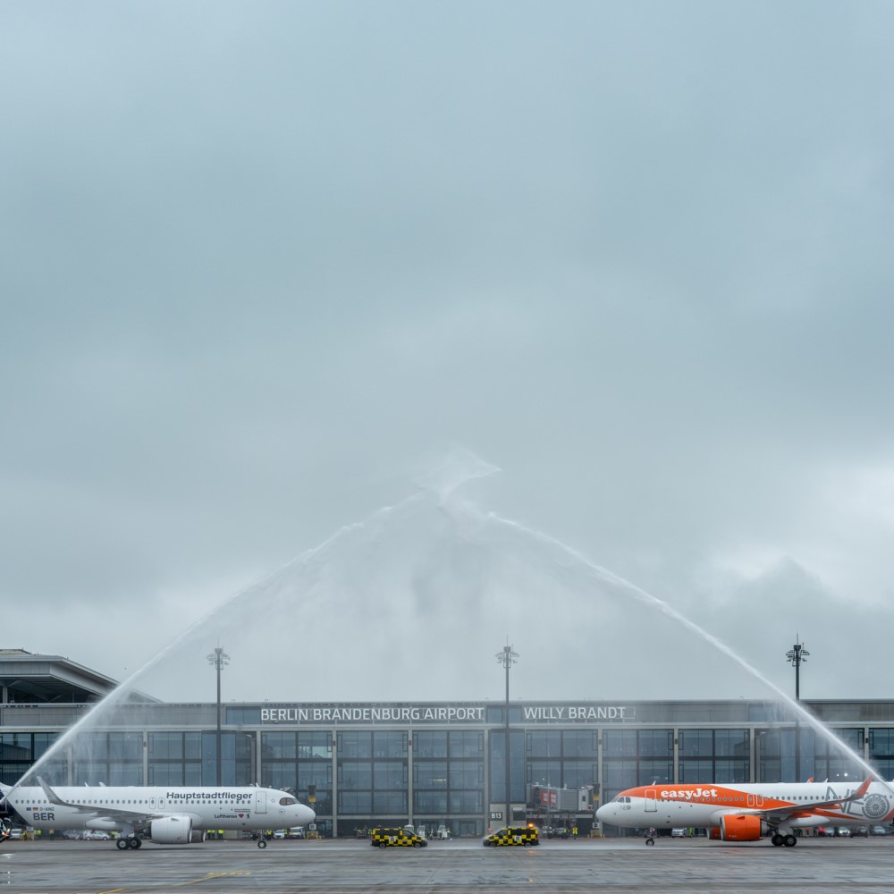 Eine Lufthansa und eine easyJet Maschine vor dem Terminal 1. Darüber zwei Wasserfontänen.