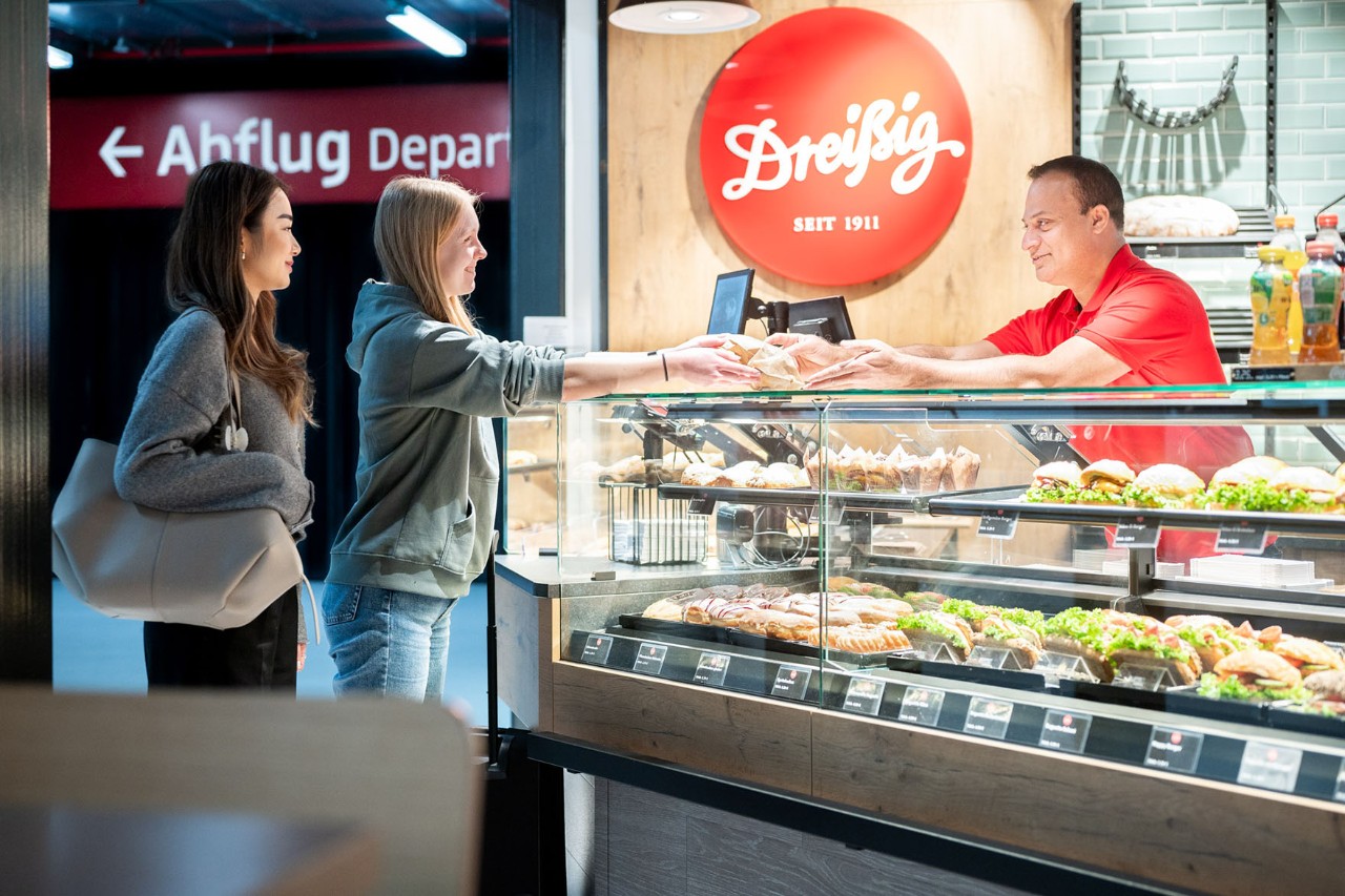 Auslage des Bäckerei-Cafés Dreißig in Terminal 2. 2 Personen werden von einer Verkaufsperson bedient.