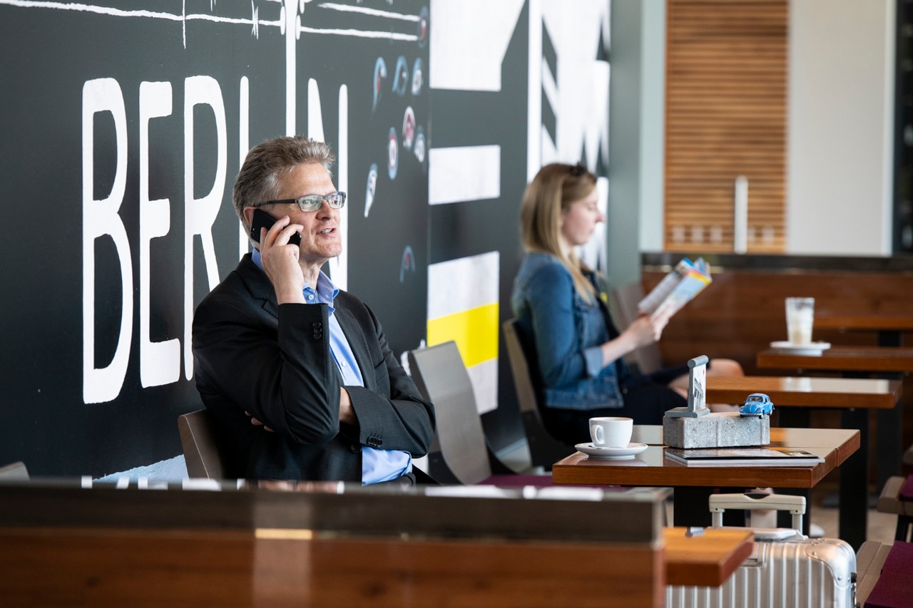2 Personen sitzen an Tischen. Eine Person telefoniert, die andere liest etwas. 