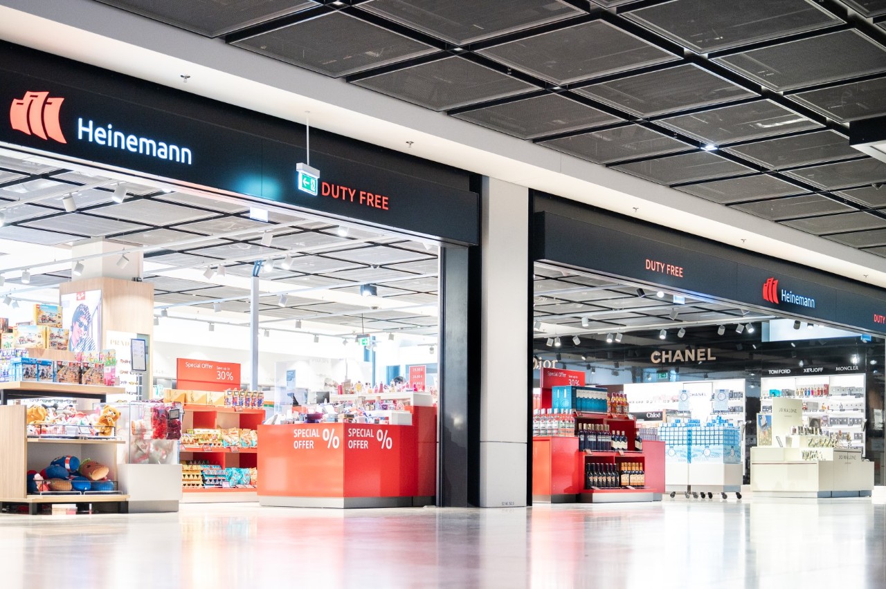 Außenansicht des Heinemann Duty Free & Travel Value Shops im Flughafen Berlin Brandenburg mit Regalen voller Produkte wie Spirituosen, Parfüm und Tabakwaren.“