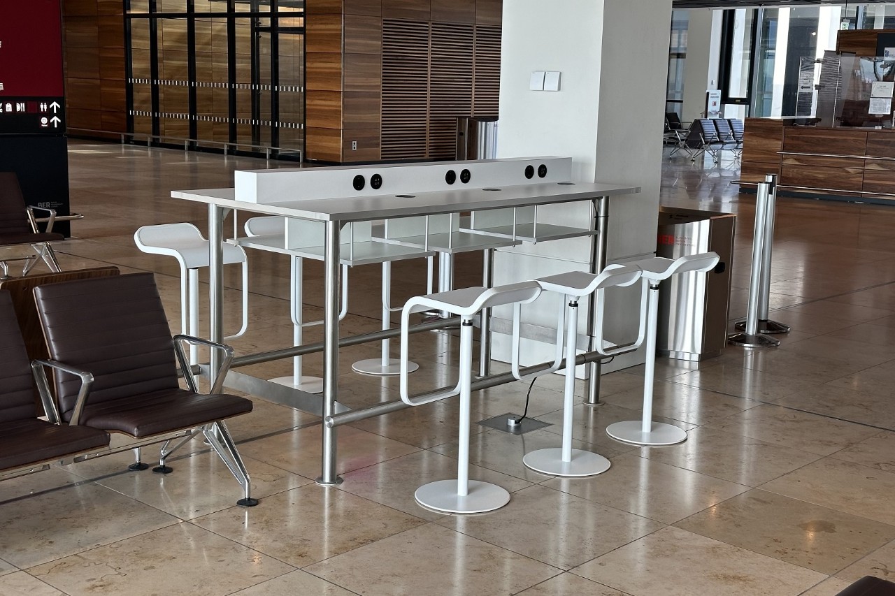 Ein Arbeitsbereich mit 6 Bistrostühlen und einem Tisch, der über Steckdosen verfügt. Im Hintergrund das Terminal. innen.