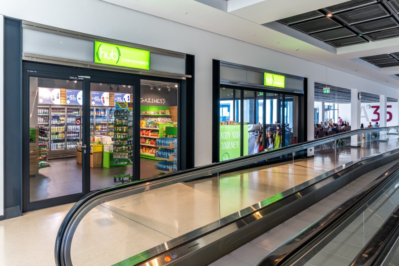 Im Vordergrund ein automatiches Rollband. Im Hintergrund der hub convenience Laden.