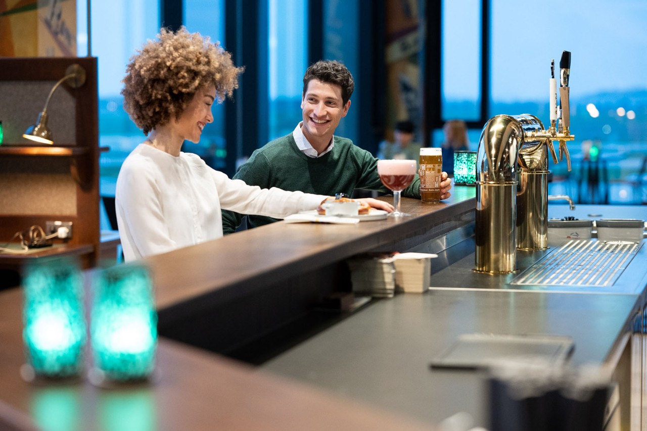 1 Mann und 1 Frau sitzen an der Bar mit Getränken auf der Theke. Dahinter das Vorfeld des BER.