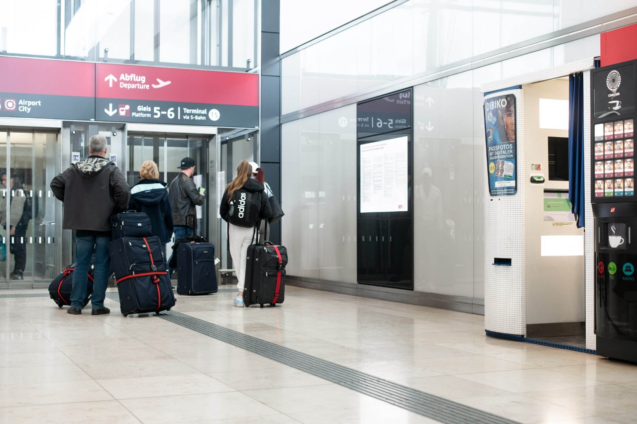 Fotoautomat. Daneben warten Reisende auf einen Fahrstuhl.