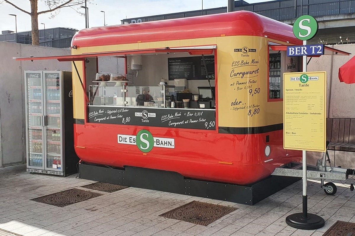 Blick auf einen Verkaufswagen, der wie eine S-Bahn aussieht. Daneben ein Getränkeautomat.