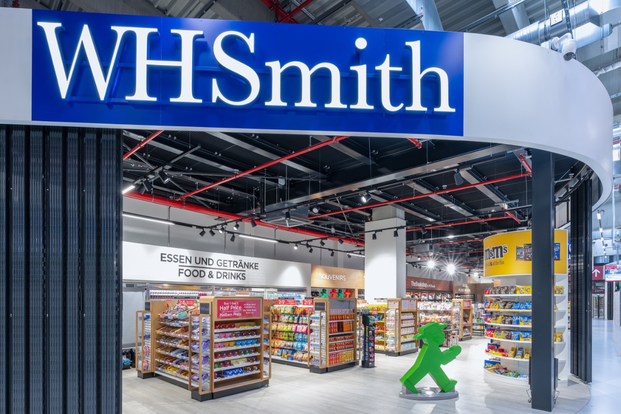 Blick von außen auf denWHSmith Laden. Ein großes Ampfelmännchen steht im Laden. Mit Essen und Getränke gefüllte Regale.