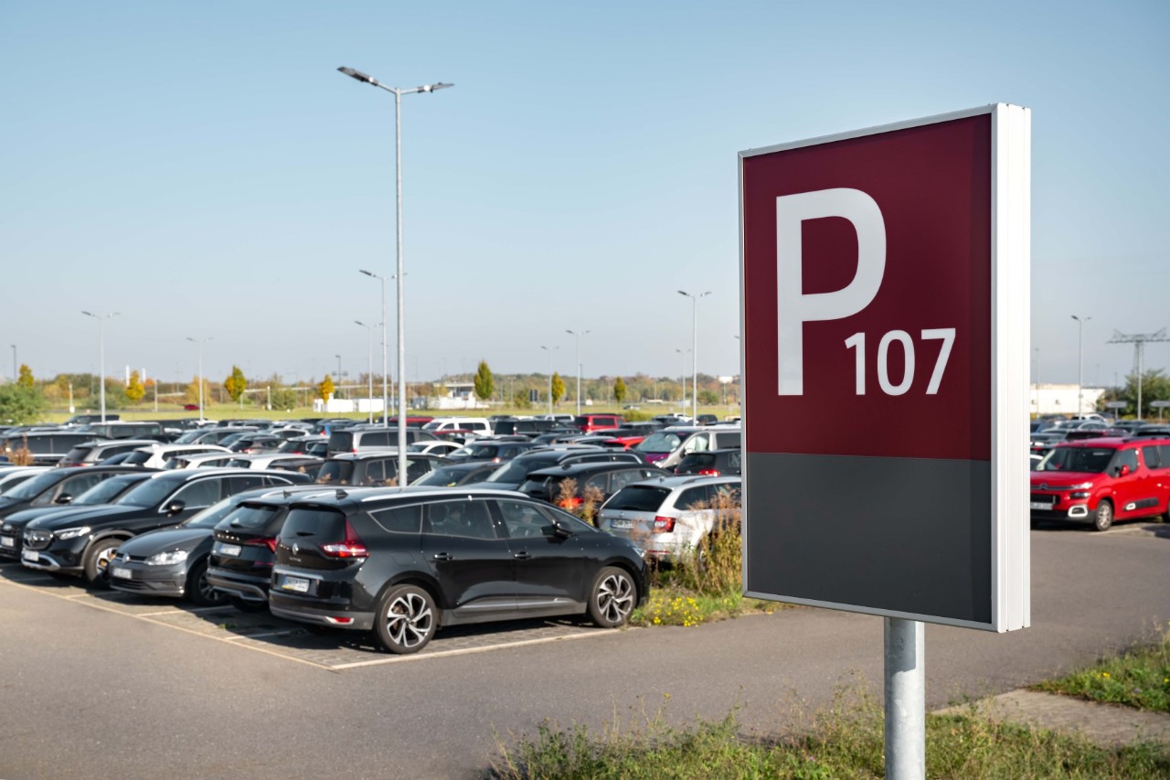 Autos auf dem Parkplatz P107. Dahinter landet ein Flugzeug. Links ein Baum.