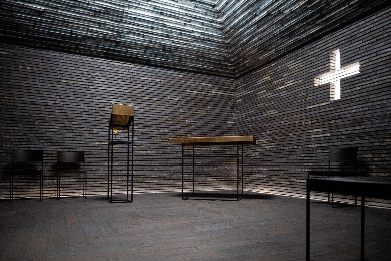 Detailaufnahme des christlichen Andachtsbereich. Von oben strahlt Licht in den mit dunklem Backstein ausgeleuchteten Raum. Ein Kreuz hängt an der Wand.