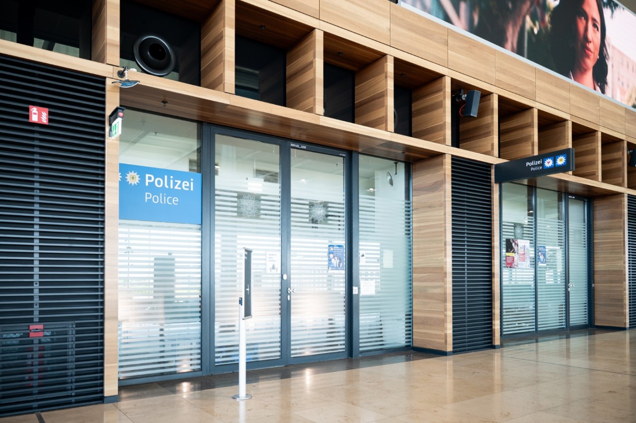 Polizeistation im Terminal 1 des Flughafens BER mit gläserner Eingangstür, darüber das Schild ‚Polizei‘ in weißer Schrift auf blauem Hintergrund.