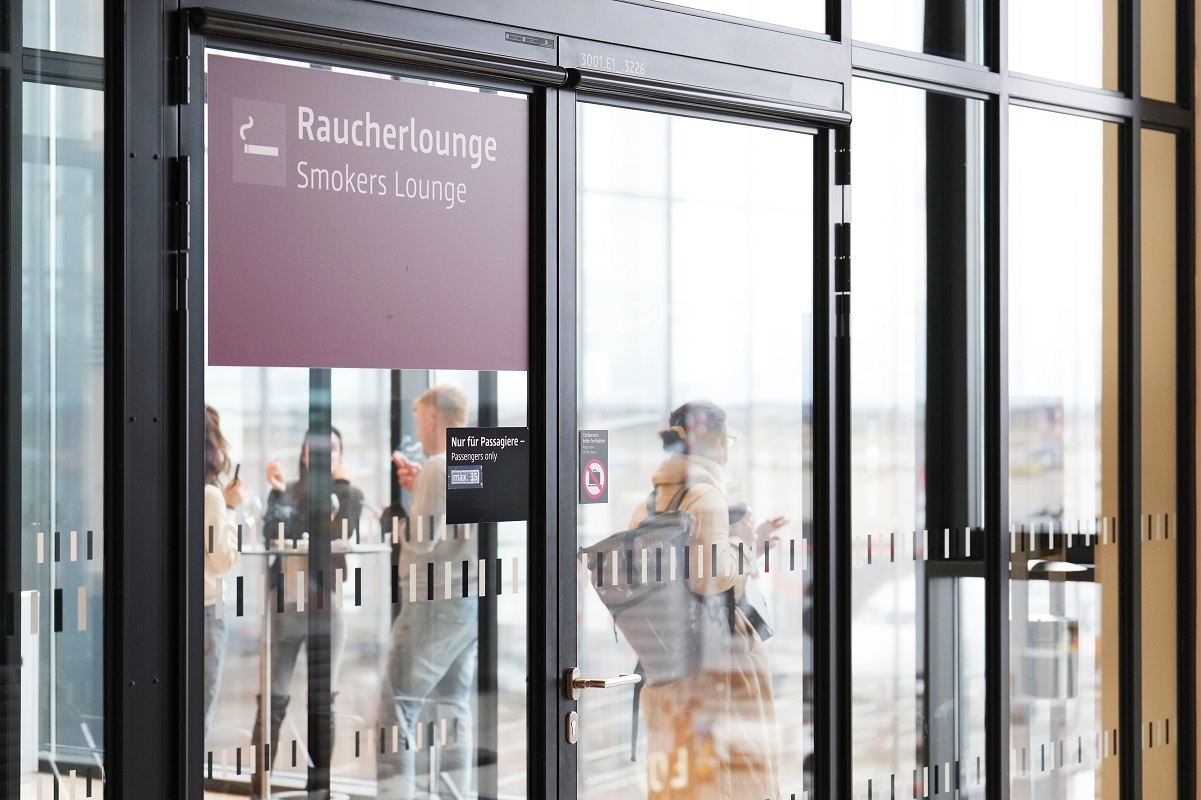 Blick in die Raucherlounge. Menschen rauchen. Im Hintergrund das Vorfeld.