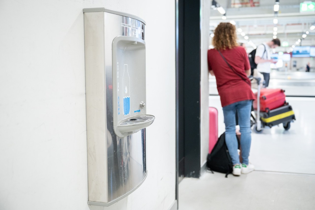 Ein Trinkwasserspender. Dahinter eine wartende Reisende, die sich an eine Wand lehnt.