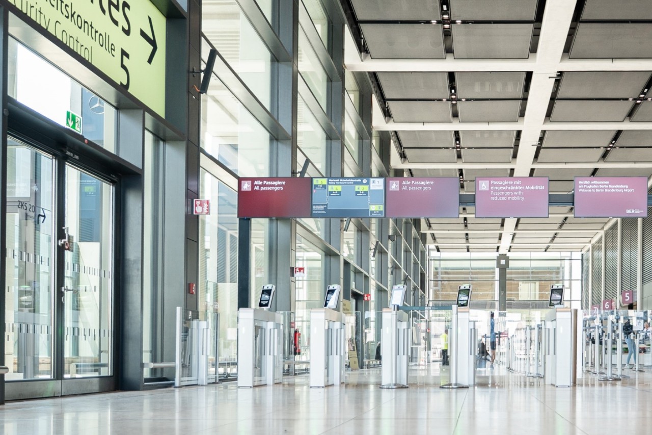 Sicherheitskontrolleschleusen in Terminal 1 des BER Darüber Monitore und eine Glastür.