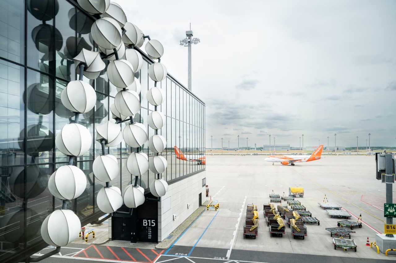 Kunstwerk Gadget: Glasfassade mit reflektierenden, weißen kugelförmigen Skulpturen, die vertikal angeordnet sind. Dahinter ein Flugzeug auf dem Vorfeld.