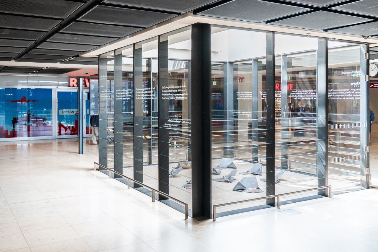 Skulptur ‚L’Albatros‘ aus neun schwebenden Papierflugzeugen im Eingangsbereich von Terminal 1 am BER. Im Hintergrund moderne Glasfassade des Terminals.