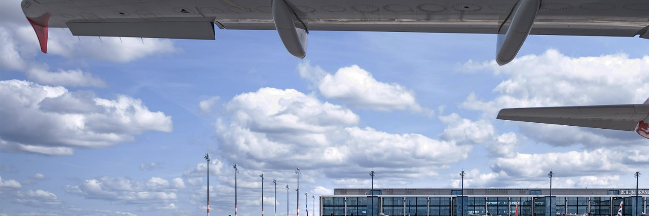 Blick unter einem Flugzeugflügel hindurch auf das obere Stück eines Terminals und den Himmel darüber