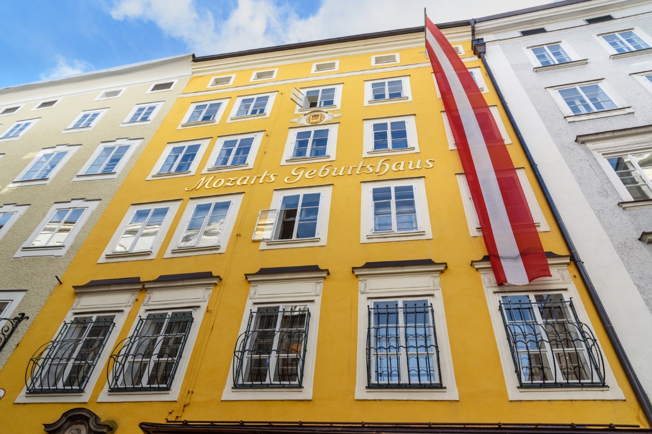 Blick auf eine Häuserfront aus der Froschperspektive. Mittig ist ein fünfstöckiges Altbau-Haus mit gelber Fassade und vielen Fenstern zu sehen. Eine langgestreckte, schmale, rote Flagge mit weißem Streifen fällt rechtsseitig des Gebäudes von oben nach unten. Links davon befinden sich ein beigefarbenes Haus und zur rechten Seite ein graues.