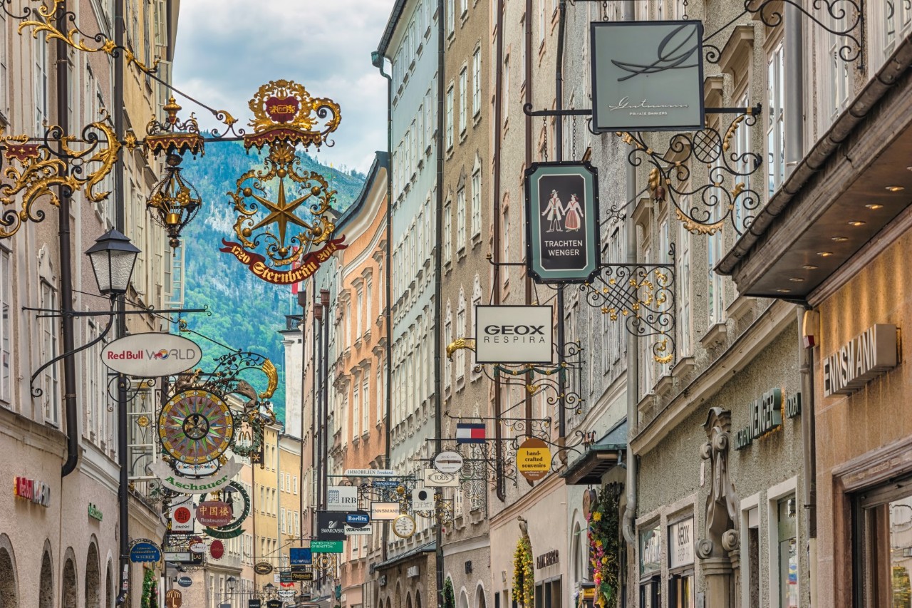 Blick in eine schmale Gasse, auf beiden Seiten Häuserfronten, die mit historischen Gewerbeschildern aus Metall verziert sind.