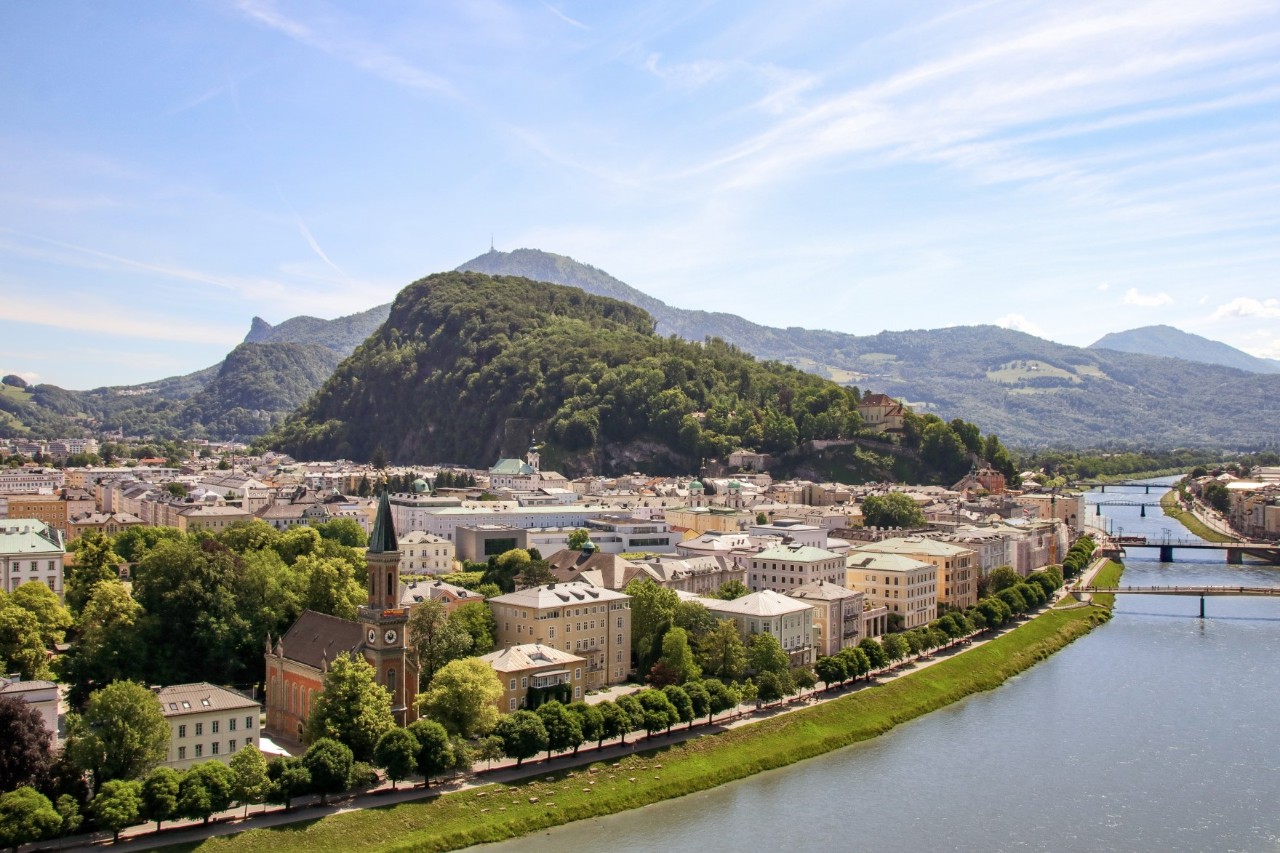 Im Zentrum des Bildes ist ein mit dichtem Wald umsäumter Berg, im Hintergrund zeichnen sich weitere bewaldete Berge ab. Rechts vorn ist ein sich schlängelnder Fluss mit 4 Brücken zu sehen. Am linken Flussufer befindet sich eine Promenade mit Baumreihe. Links des Flusses ist eine dicht bebaute Stadt erkennbar.