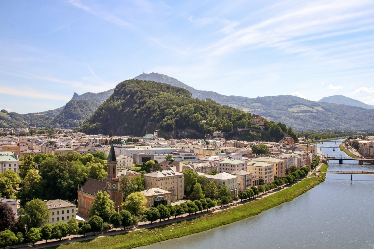 Dichter mit Wald umsäumter Berg, im Hintergrund zeichnen sich weitere bewaldete Berge ab. Rechts vorn ist ein sich schlängelnder Fluss mit 4 Brücken zu sehen. Am linken Flussufer befindet sich eine Promenade mit Baumreihe. Links des Flusses ist eine dicht bebaute Stadt erkennbar.