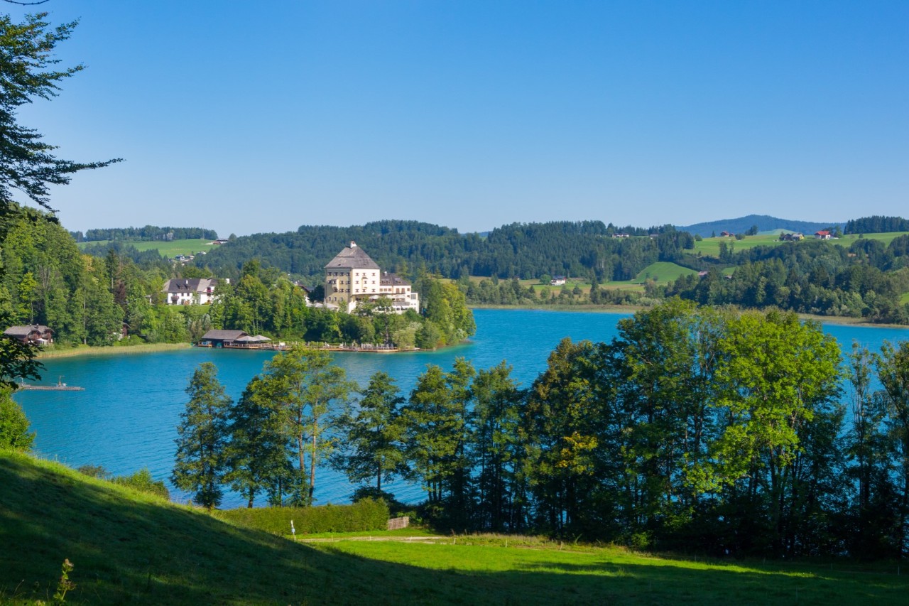 Blick von einer hügeligen grünen Landschaft auf einen See mit kristallblauem Wasser. Der See ist in eine bergige Landschaft mit Wäldern, grünen Wiesen und einzelnen Häusern eingebettet.