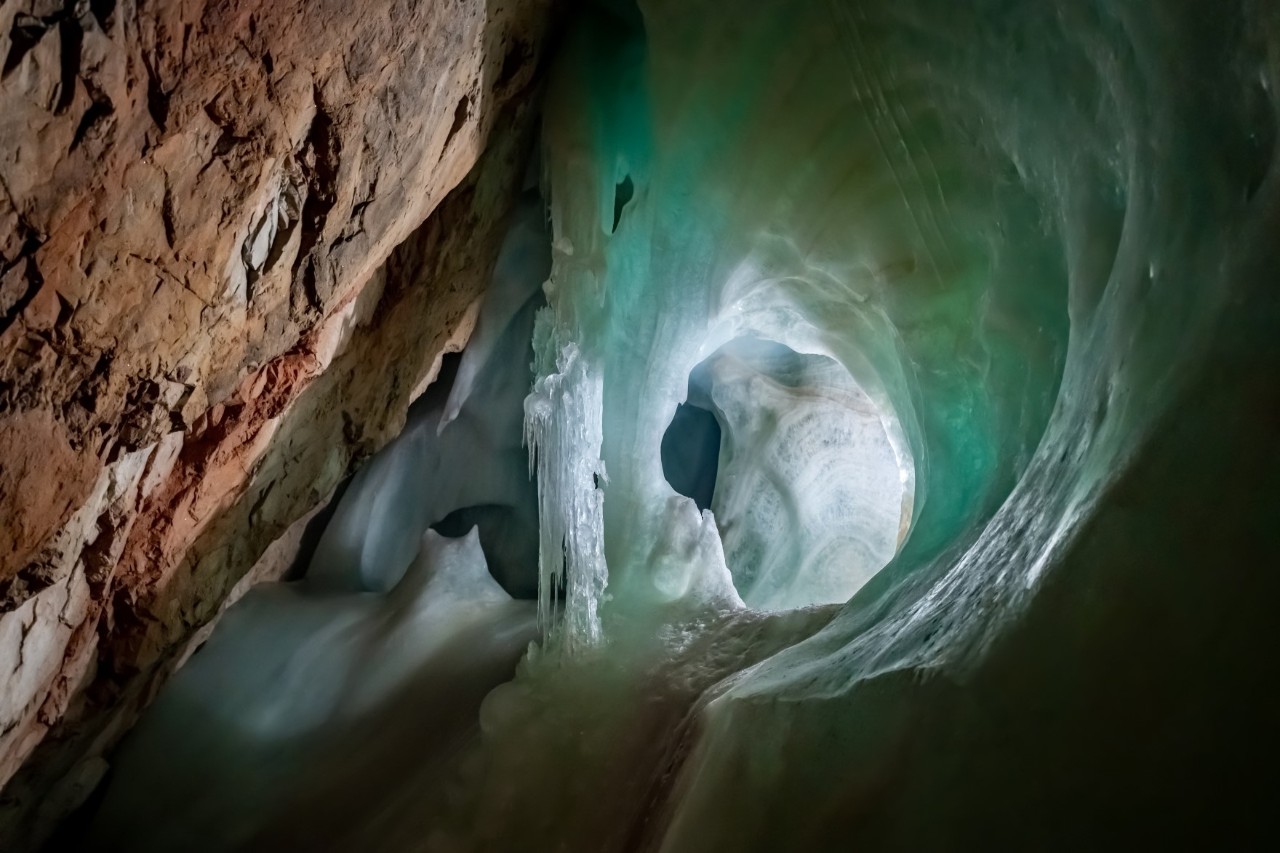 Blick in eine dunkle Höhle aus braunem Gestein und weiß-türkisleuchtendem Eisformationen.