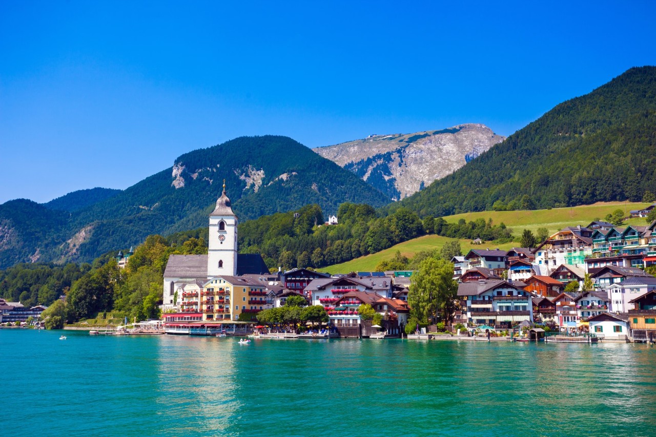 Blick von einem See auf eine am Ufer gelegene Ortschaft. Dahinter erstreckt sich eine grüne Wiese in Hanglage und bewaldete Berge.