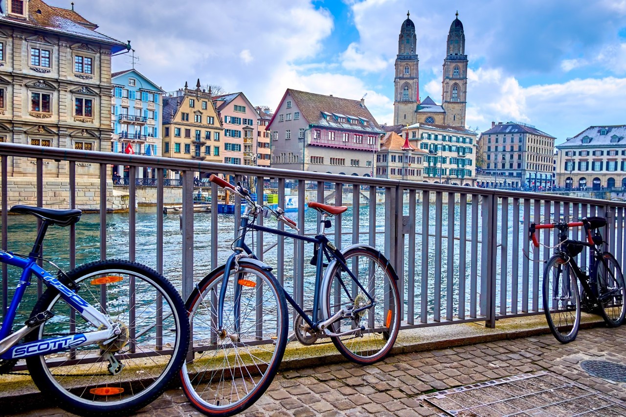 Blick auf drei Fahrräder, die an einem Geländer abgestellt sind, hinter dem ein Fluss verläuft. Auf der gegenüberliegende Flussseite sind mehrere Stadthäuser in verschiedenen Farben und eine imposante Kirche mit zwei hohen Türmen zu sehen.