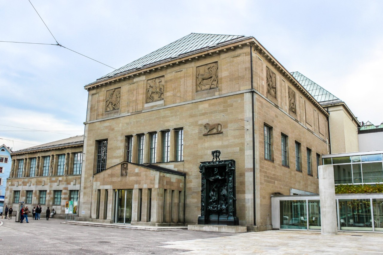 Blick auf ein wuchtiges Steingebäude mit großen Fenstern.  Die oberen Fenster sind mit Steinbildern zugemauert. Auf der rechten Seite befindet sich ein kleiner, moderner Anbau mit verglaster Fassade. Links vom Haupthaus verläuft ein alter Seitenflügel.