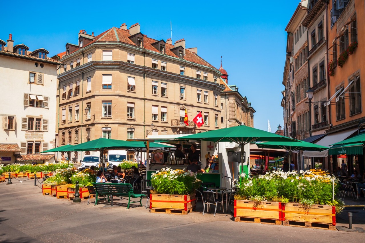 Blick auf einen Platz, der von vierstöckigen Wohnhäusern umgeben ist und in dessen Mittelpunkt sich ein Straßencafé befindet. Das Café ist von bepflanzten Kästen begrenzt. Die Tische und Stühle sind mit grünen Sonnenschirmen überdeckt.