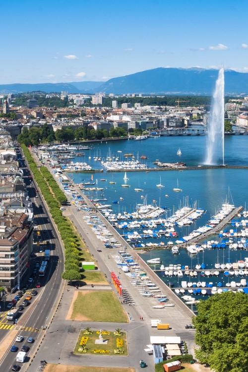 Luftaufnahme einer Stadt, die direkt an einem grün-blauen See liegt, in dem sich ein Hafen mit vielen kleinen Booten und eine Wasserfontäne befindet. Im Hintergrund sind flache Berge zu sehen. Entlang des Ufers verläuft eine Straße, deren Straßenränder mit Bäumen bepflanzt sind.