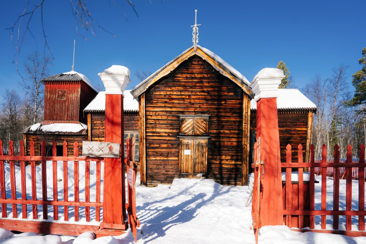 Eine Holzkirche mit rotem Zaun im Schnee, vor blauem Himmel, umgeben von einigen Bäumen.