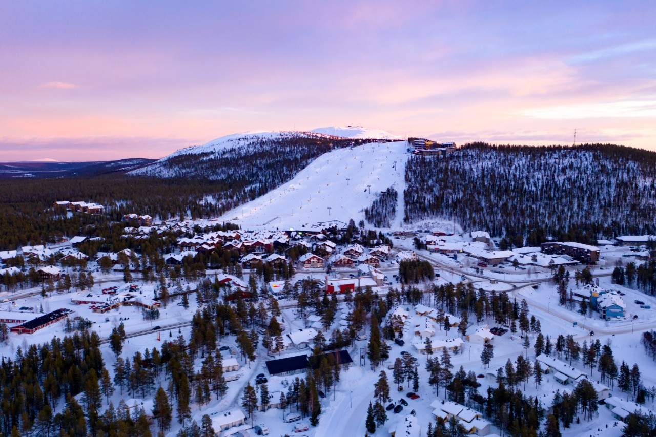 Ein Skigebiet im Winter mit einer Skipiste und verschneiten Häusern, umgeben von Wald. Der Himmel ist in pastellrosa und lila Tönen gefärbt.