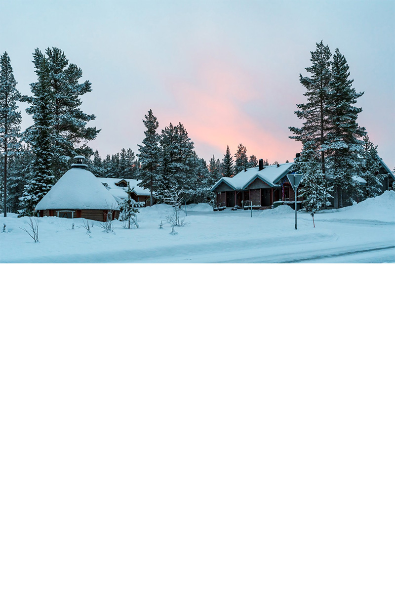 Verschneite Holzhäuser in einem winterlichen Wald mit einem teils rosa gefärbten Himmel und schneebedeckten Bäumen. 