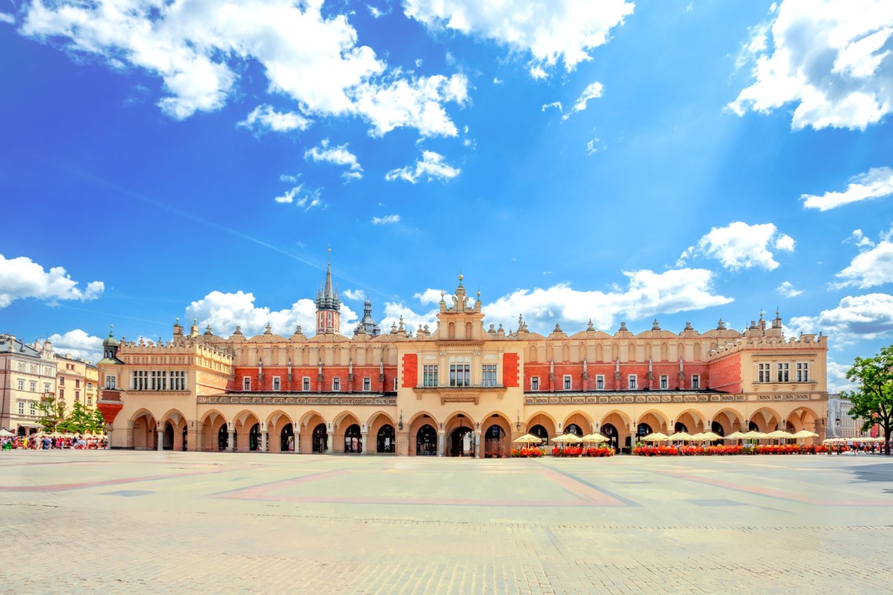 Längsseite eines großen Gebäudes mit neugotischen Arkaden und Säulenkapitellen vor blauem Himmel, davor ist ein großer menschenleerer Marktplatz sichtbar 