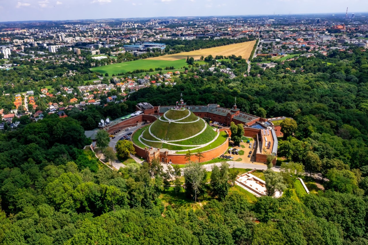Luftaufnahme eines Hügels, der von Gebäuden und Wald umgeben ist. Am Horizont ist die Stadt zu sehen 