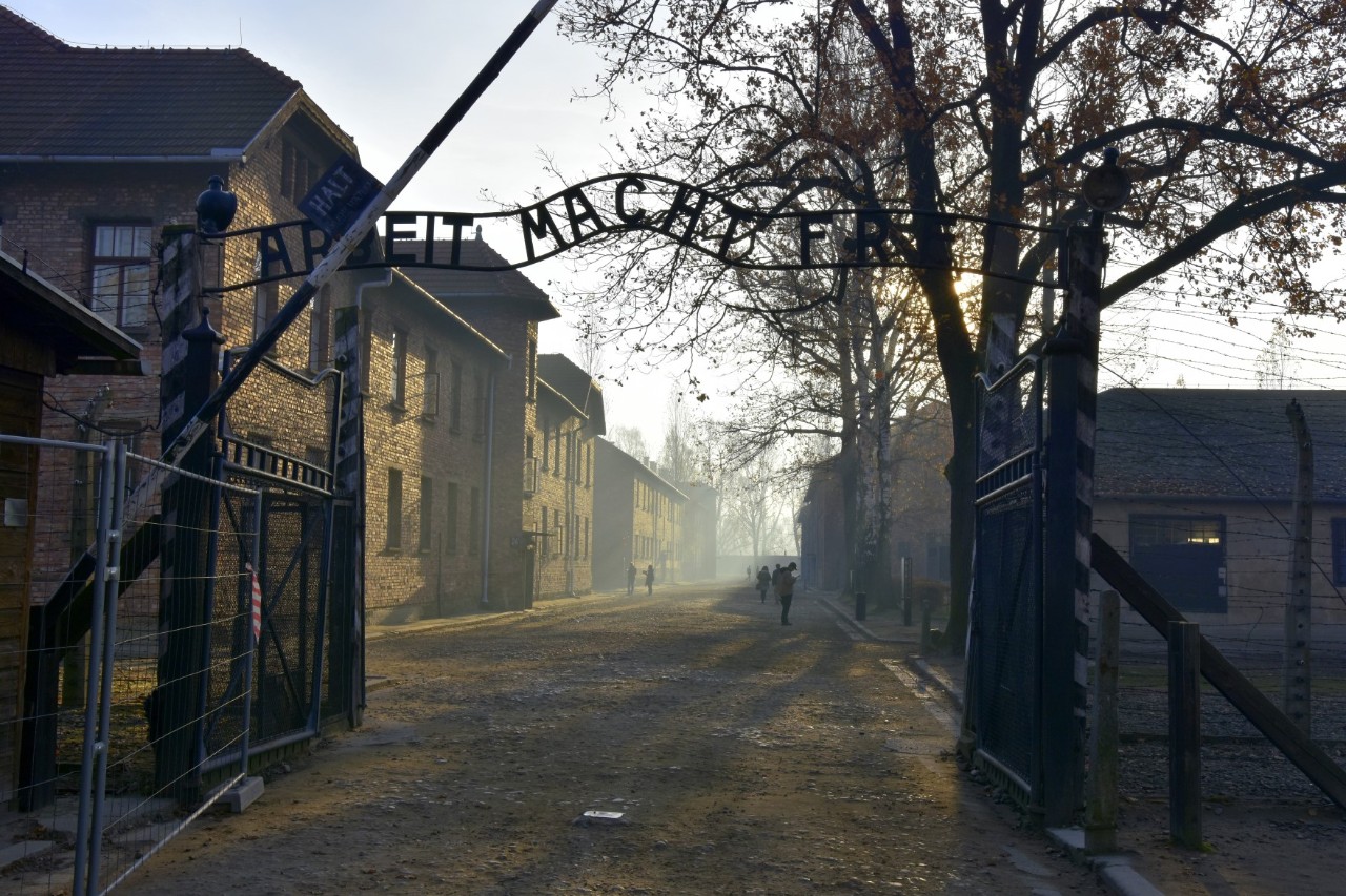 Blick auf das Eingangstor zum Konzentrationslager mit der Aufschrift „Arbeit macht frei“. Die Schranke zum Eingang steht schräg nach oben offen, eine Straße führt ins Lager hinein, links ist eine Häuserreihe zu sehen, rechts Bäume und weitere Gebäude 