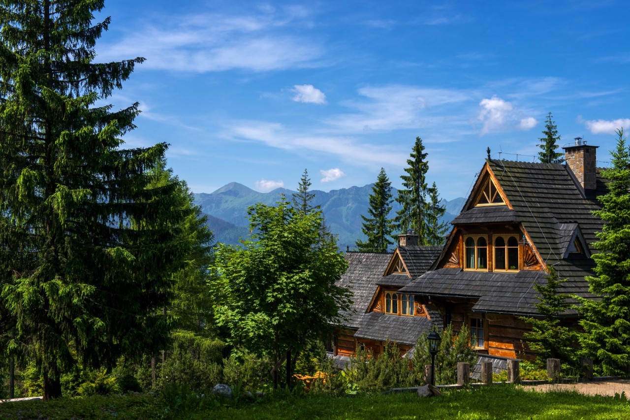 Blick auf traditionelle Holzhäuser in den Tatra-Bergen. Die Häuser sind umrahmt von Nadelbäumen, am Horizont zeichnet sich eine Berglandschaft ab 