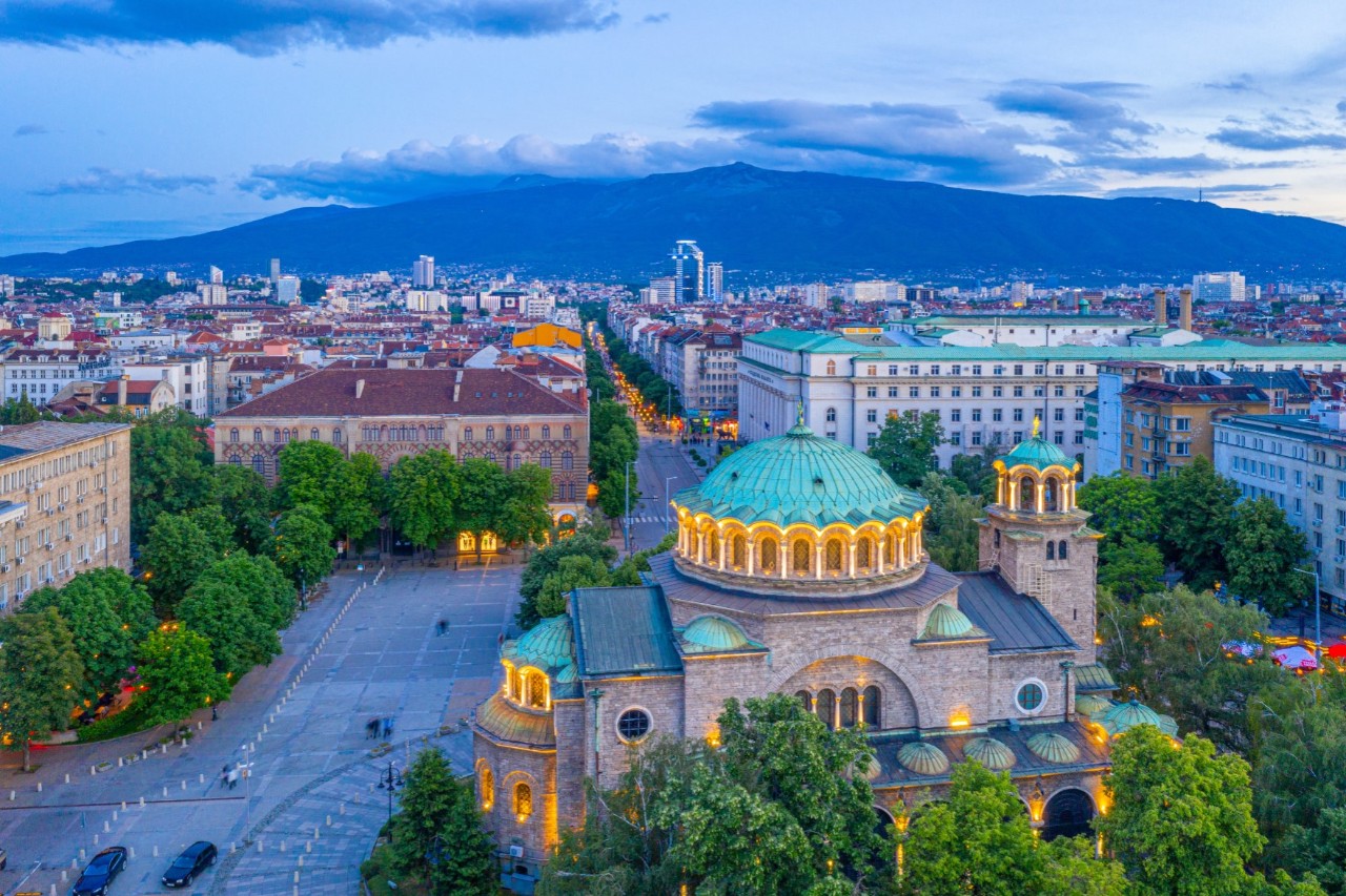 Das Bild zeigt eine Stadtansicht in der Dämmerung. Im Vordergrund steht eine große, beleuchtete Kirche mit einer markanten grünen Kuppel und mehreren kleinen grünen Kuppeln und Halbkuppeln. Die Kirche ist umgeben von Bäumen und einem menschenleeren Platz. Im Hintergrund sind erst Häuser, dann in weiter Ferne ein Gebirge zu sehen.