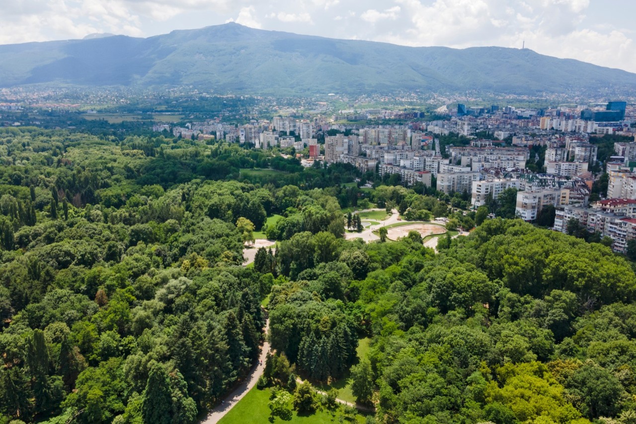 Auf dem Bild ist im Vordergrund ein großer, dicht bewaldeter Park zu sehen, der an eine Stadtlandschaft mit zahlreichen Wohngebäuden auf der rechten Bildseite grenzt. Im Hintergrund von Park und Häusern ist ein Gebirge zu sehen.