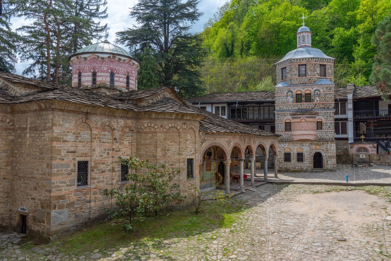 Abgebildet ist ein historisches Kloster, das in einer grünen Umgebung liegt. Die Gebäude sind aus Naturstein gebaut und haben leicht gewölbte Dächer, Kuppeln und verzierte Eingänge mit Rundbögen. Davor ist ein kopfsteingepflasterter Platz zu sehen.