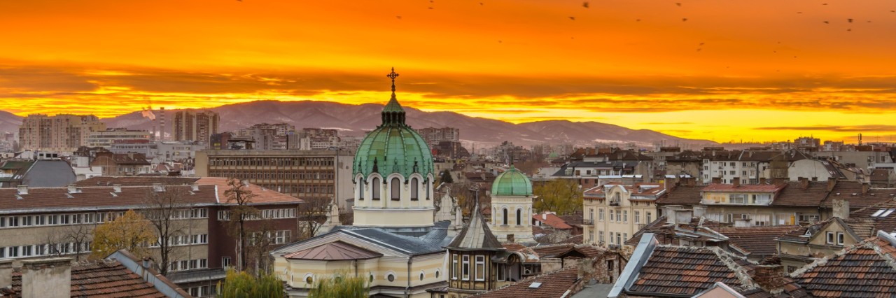 Das Bild zeigt den Blick über die Dächer einer europäischen Stadt mit bürgerlichen Altbauten bei Sonnenuntergang. Im Mittelpunkt ist eine Kirche mit einer großen und einer kleineren grünen Kuppel zu sehen. Am Horizont erstrecken sich Berge, hinter denen die Sonne langsam untergeht. Der Himmel ist oben orange und zum Horizont hin gelb.