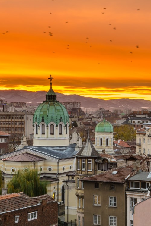 Das Bild zeigt den Blick über die Dächer einer europäischen Stadt mit bürgerlichen Altbauten bei Sonnenuntergang. Im Mittelpunkt ist eine Kirche mit einer großen und einer kleineren grünen Kuppel zu sehen. Am Horizont erstrecken sich Berge, hinter denen die Sonne langsam untergeht. Der Himmel ist oben orange und zum Horizont hin gelb.