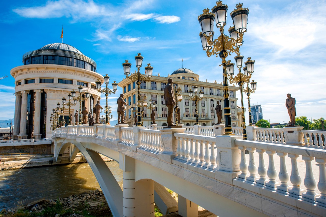 Blick auf eine weiße Bogenbrücke, auf der mehrere Straßenlaternen und Statuen in Menschenform zu sehen sind. Die Brücke läuft auf zwei markante Gebäude zu. Links ist ein rundes Haus und rechts ein größeres, rechteckiges Haus, beide in Beigetönen, zu sehen.