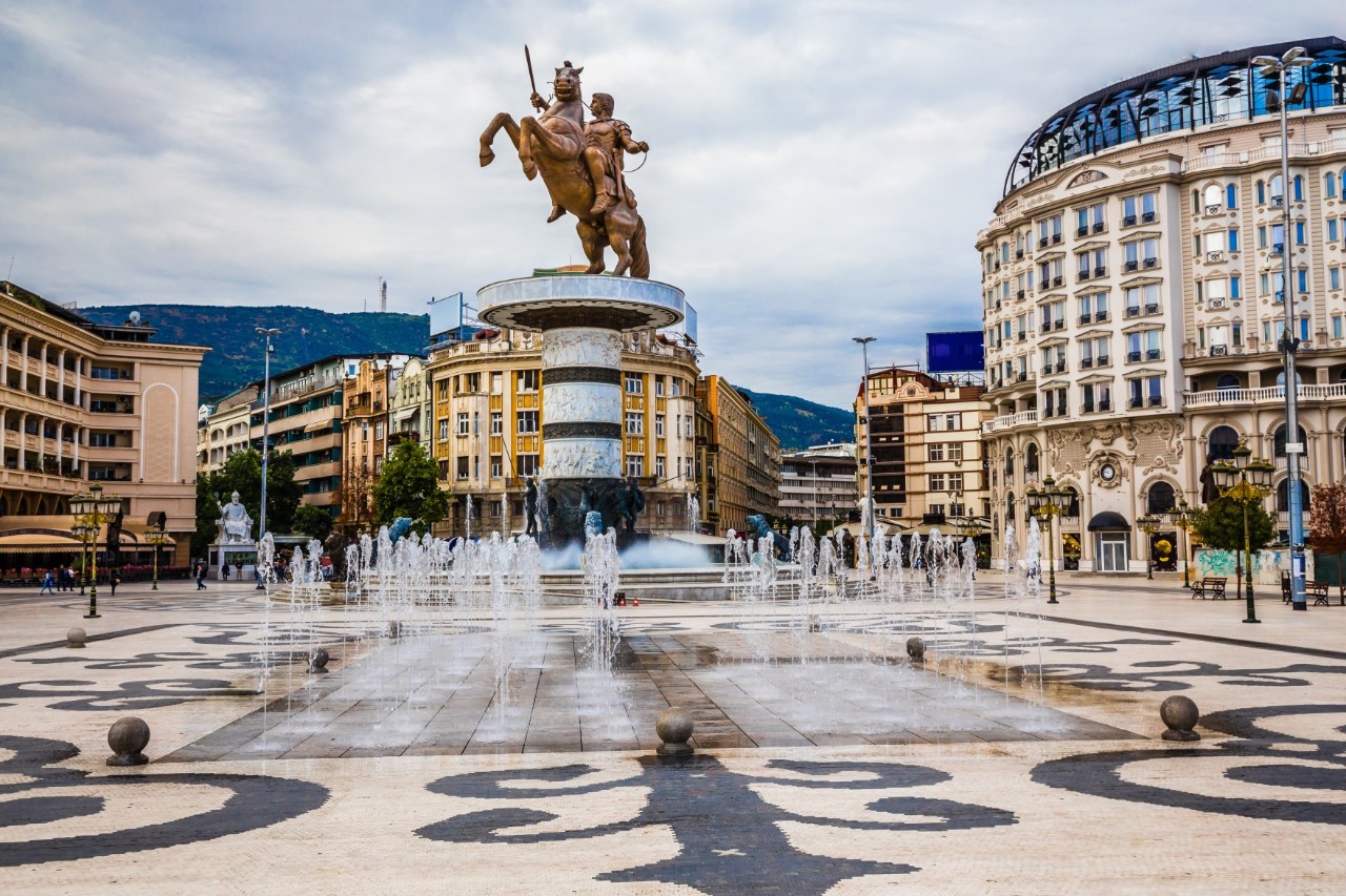 Blick auf einen gepflegten, städtischen Platz, in dessen Mitte eine goldene Reiterstatue auf einem Sockel steht. Davor sind kleine Wasserfontänen und ein heller Boden mit schwarzen Ornamenten zu sehen. Der Platz ist von mehrgeschossigen, teilweise runden Häuser umgeben.