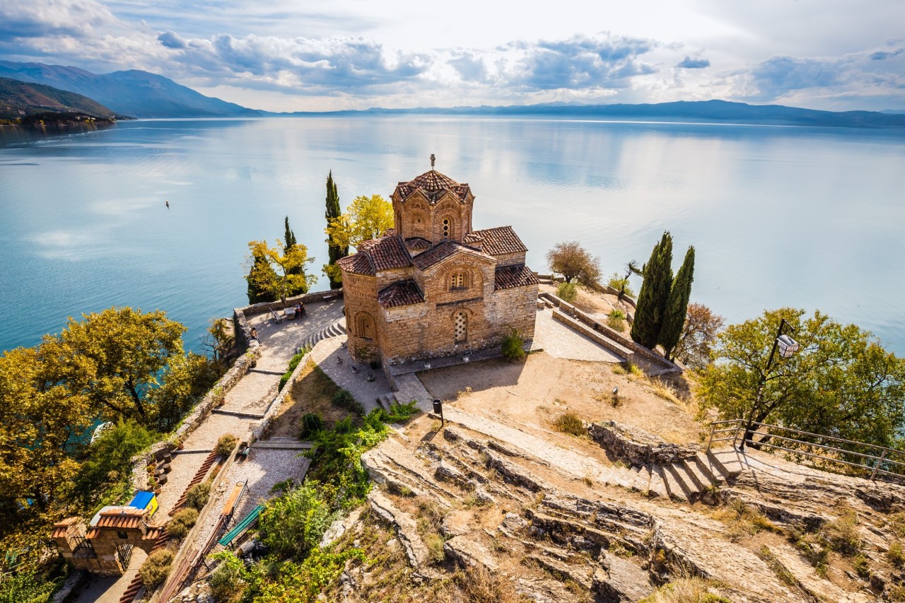 Blick auf eine orthodoxe Kirche, die auf einer Landzunge an einem mit flachen Bergen umsäumten See steht. Die Kirche ist von grünen Bäumen und diversen Steinformationen umgeben.